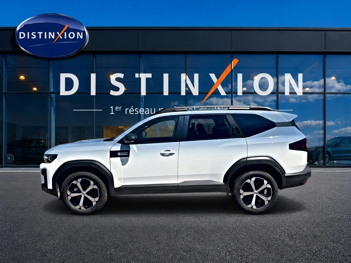 Vue de profil côté gauche d'un Dacia Bigster blanc avec jantes alu et barres de toit devant une vitrine Distinxion.