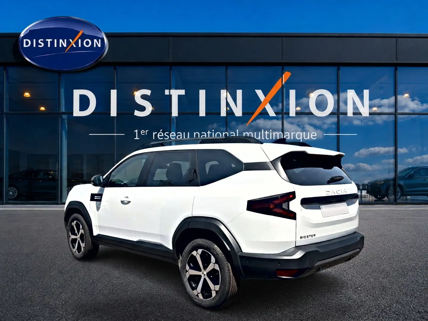 Vue 3/4 arrière droite du Dacia Bigster blanc avec jantes alu et feux arrière en forme de Z devant une vitrine Distinxion.
