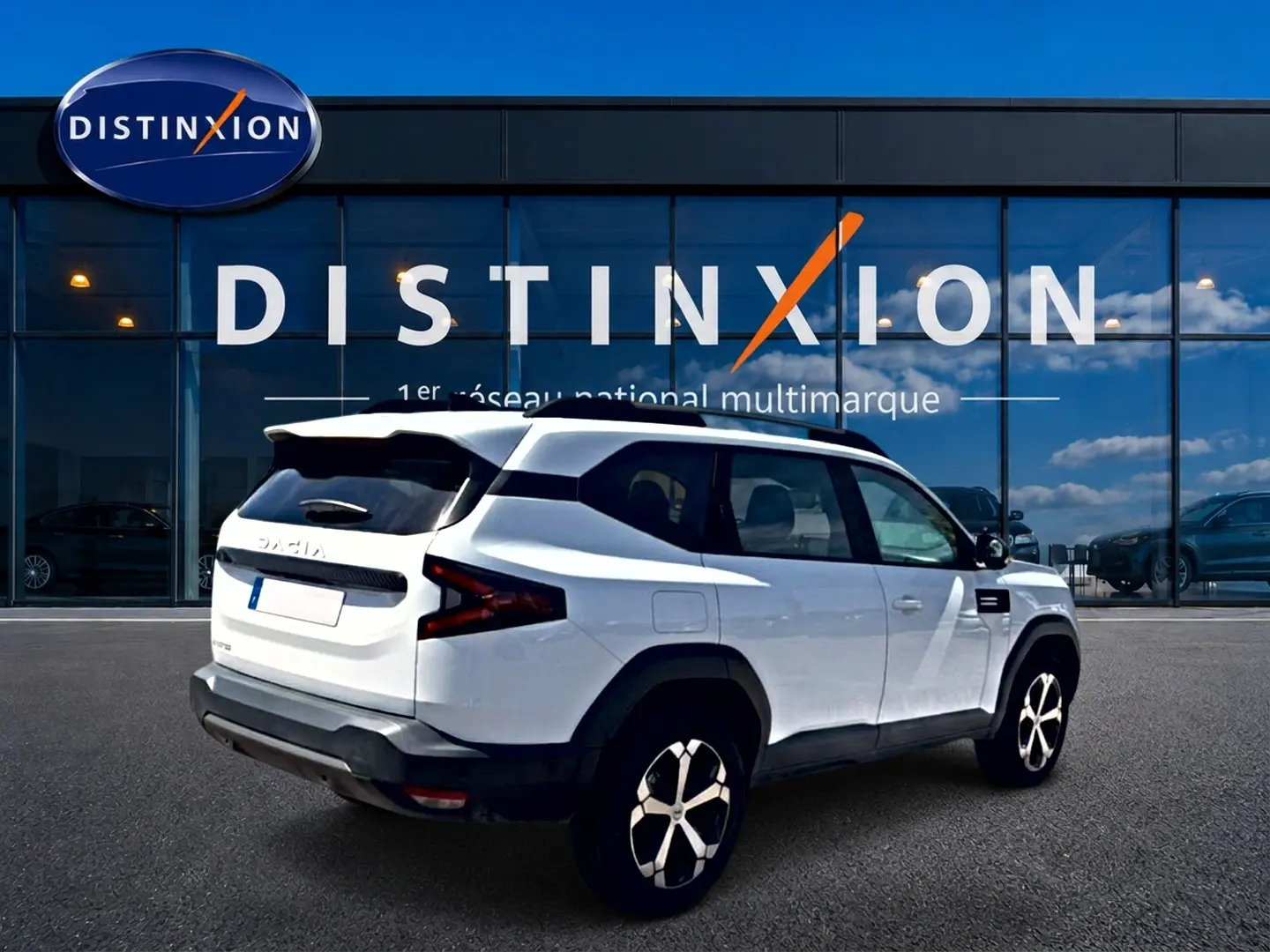 Vue 3/4 arrière droite du SUV blanc Dacia Bigster 2026 avec jantes alu et barres de toit noires devant un bâtiment Distinxion.