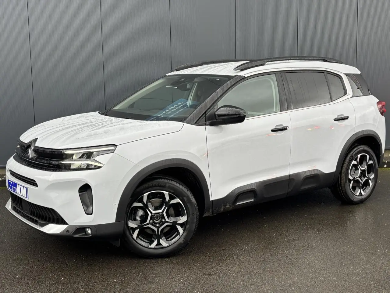Vue 3/4 avant droite d’un Citroën C5 Aircross blanc Okénite avec jantes alliage bi-ton et barres de toit noires.
