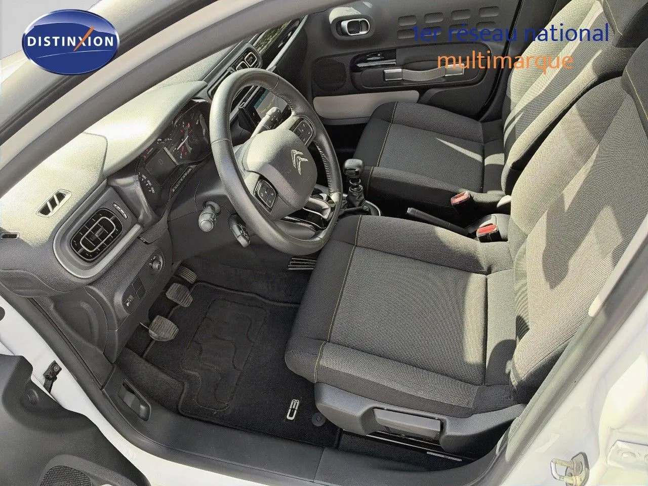 Vue intérieure côté conducteur de la Citroën C3 blanche 2021, montrant le volant, les sièges tissu gris et la boîte manuelle.