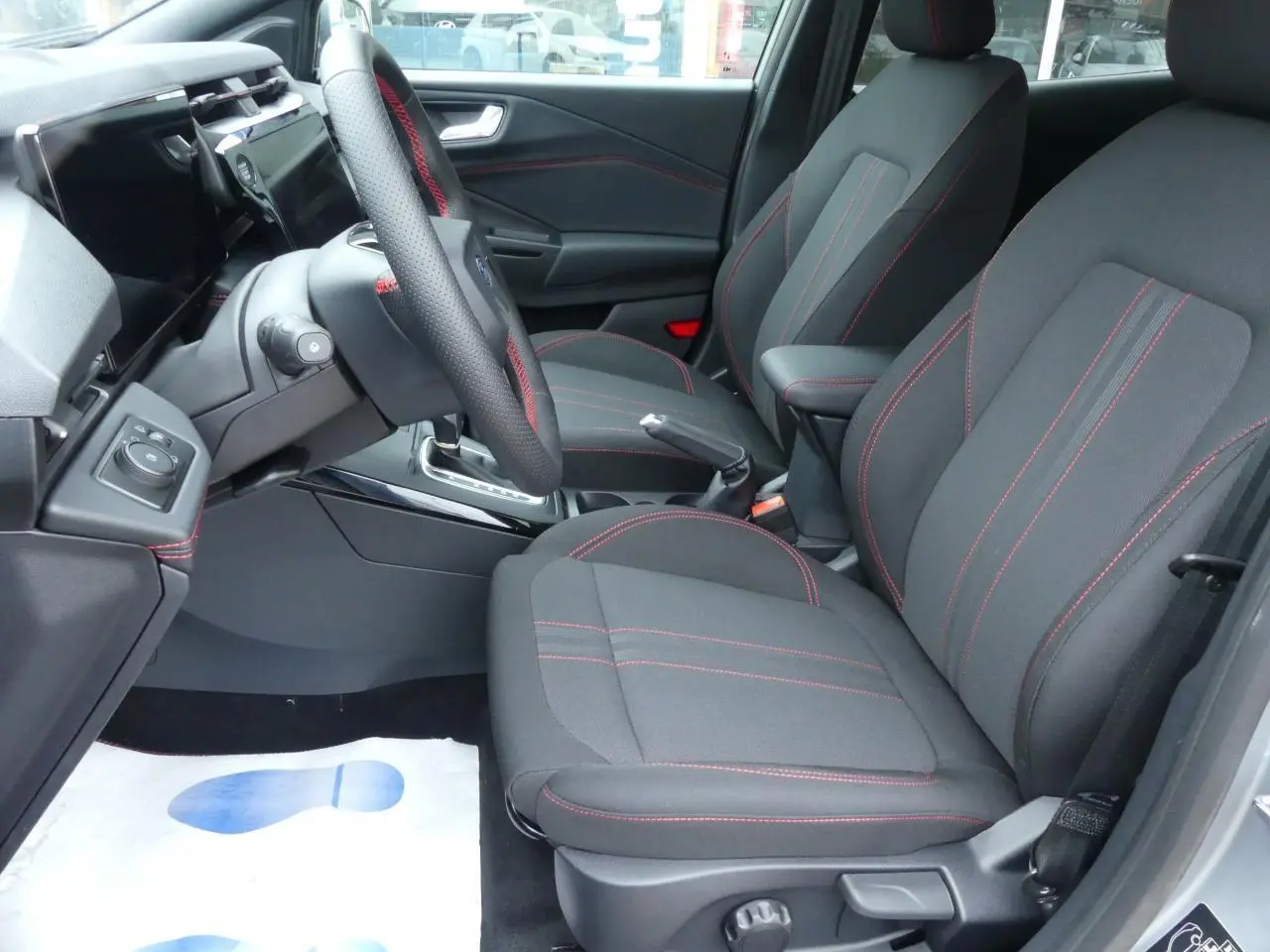 Intérieur avant droit du Ford Puma 2025 avec sièges tissu noirs et surpiqûres rouges, volant cuir perforé.