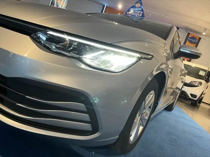 Vue 3/4 avant droit d'une Volkswagen Golf 8 argent métallisé avec phare allumé en intérieur showroom.