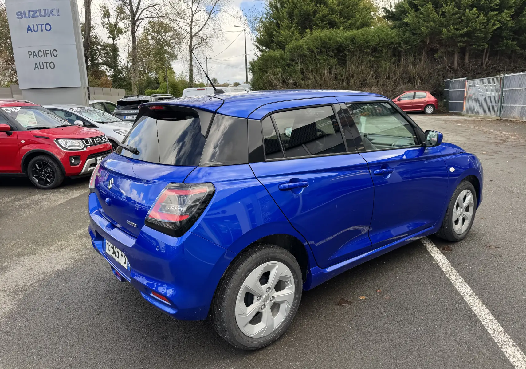 Vue 3/4 arrière droite d'une Suzuki Swift bleu électrique avec toit noir, garée sur un parking extérieur.