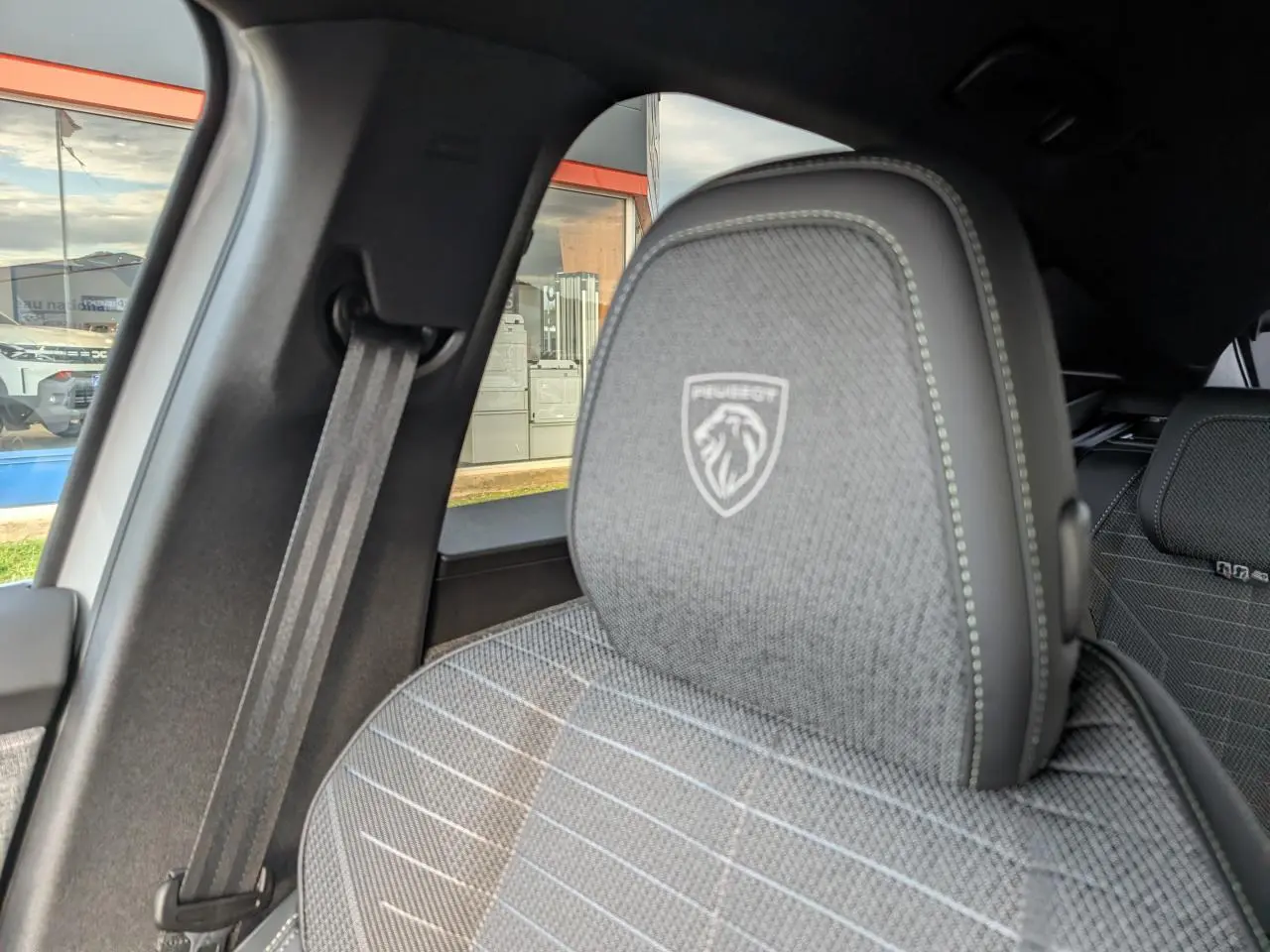 Gros plan sur l'appui-tête avant gauche gris avec logo Peugeot, intérieur tissu et ceinture de sécurité visible.