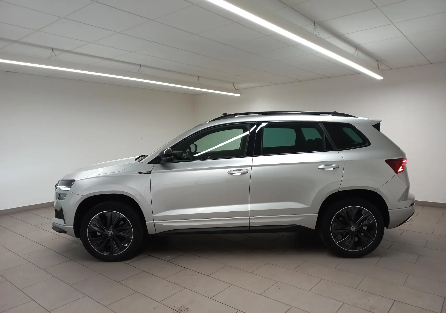 Vue de profil côté gauche d’un Skoda Karoq gris argent 2025 avec toit panoramique et jantes noires en intérieur neutre.