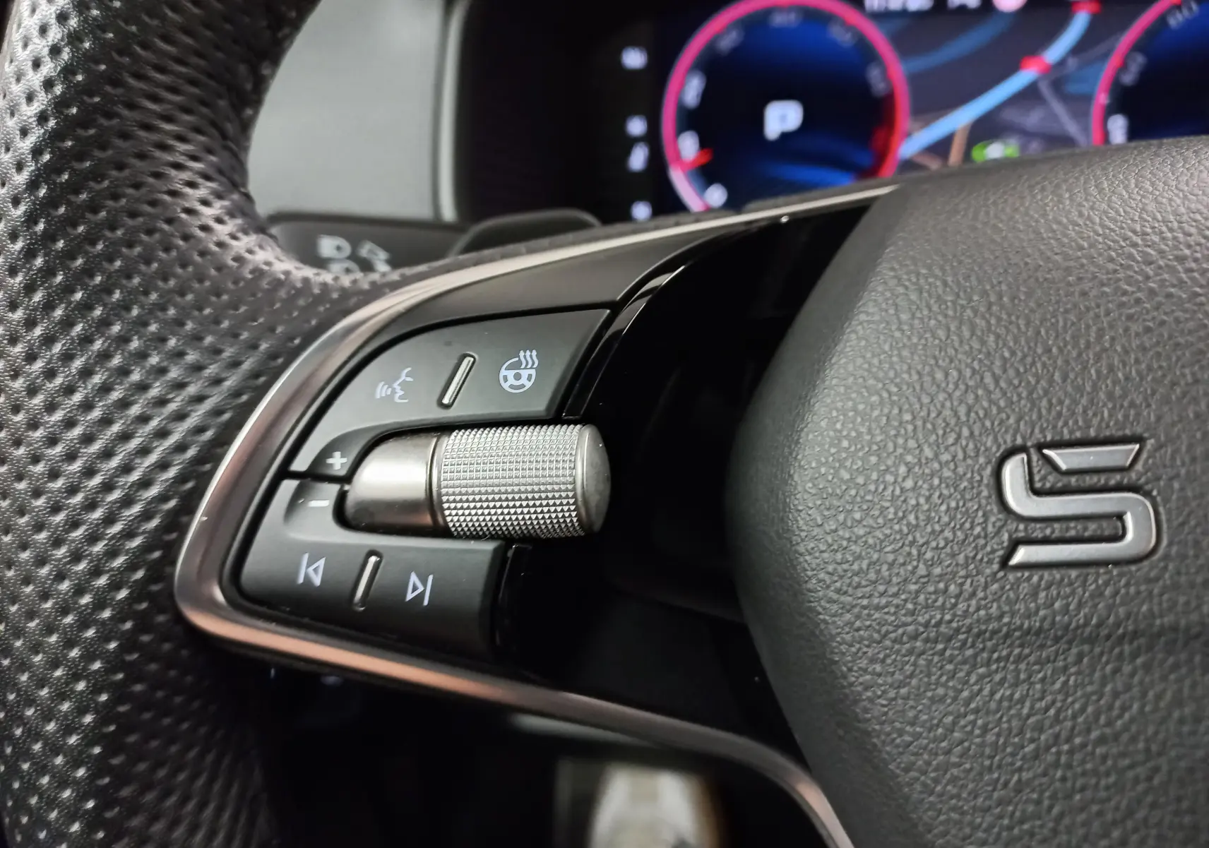 Gros plan sur les commandes gauche du volant perforé du Skoda Karoq gris argent 2025 avec molette métallique et affichage numérique en arrière-plan.