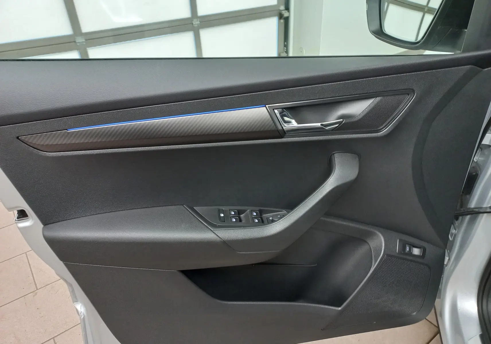Intérieur côté gauche de la porte avant gris argent du Skoda Karoq 2025 avec insert carbone et éclairage bleu subtil.