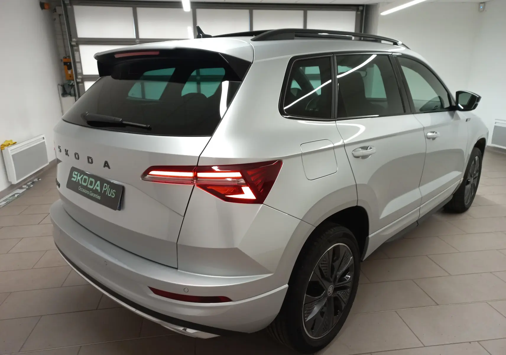 Vue 3/4 arrière droite du Skoda Karoq gris argent 2025 avec toit panoramique et feux arrière LED allumés.