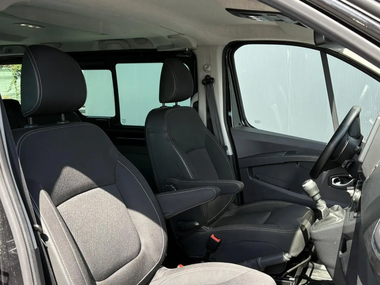 Vue intérieure côté conducteur du Nissan Primastar Combi noir Minuit, montrant sièges avant avec accoudoirs et levier de vitesse manuel.