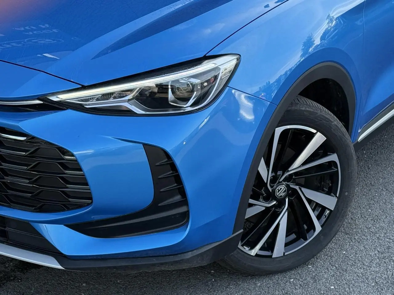 Gros plan sur l’avant droit bleu Como du MG ZS 1.5 Hybrid+ 197 Luxury avec phare LED et jante alliage 18 pouces.