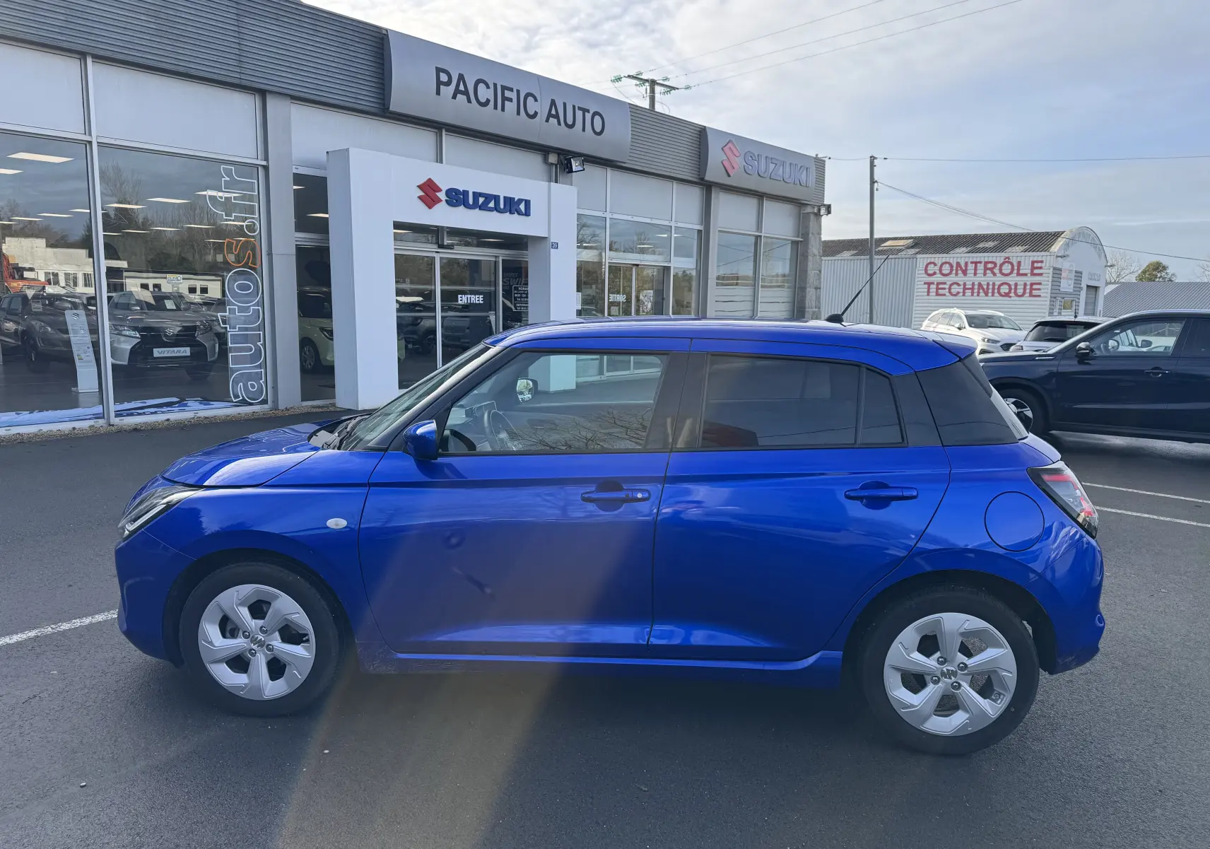 Profil côté gauche d'une Suzuki Swift 1.2 Hybrid Privilege bleu brillant, garée devant un concessionnaire Suzuki.