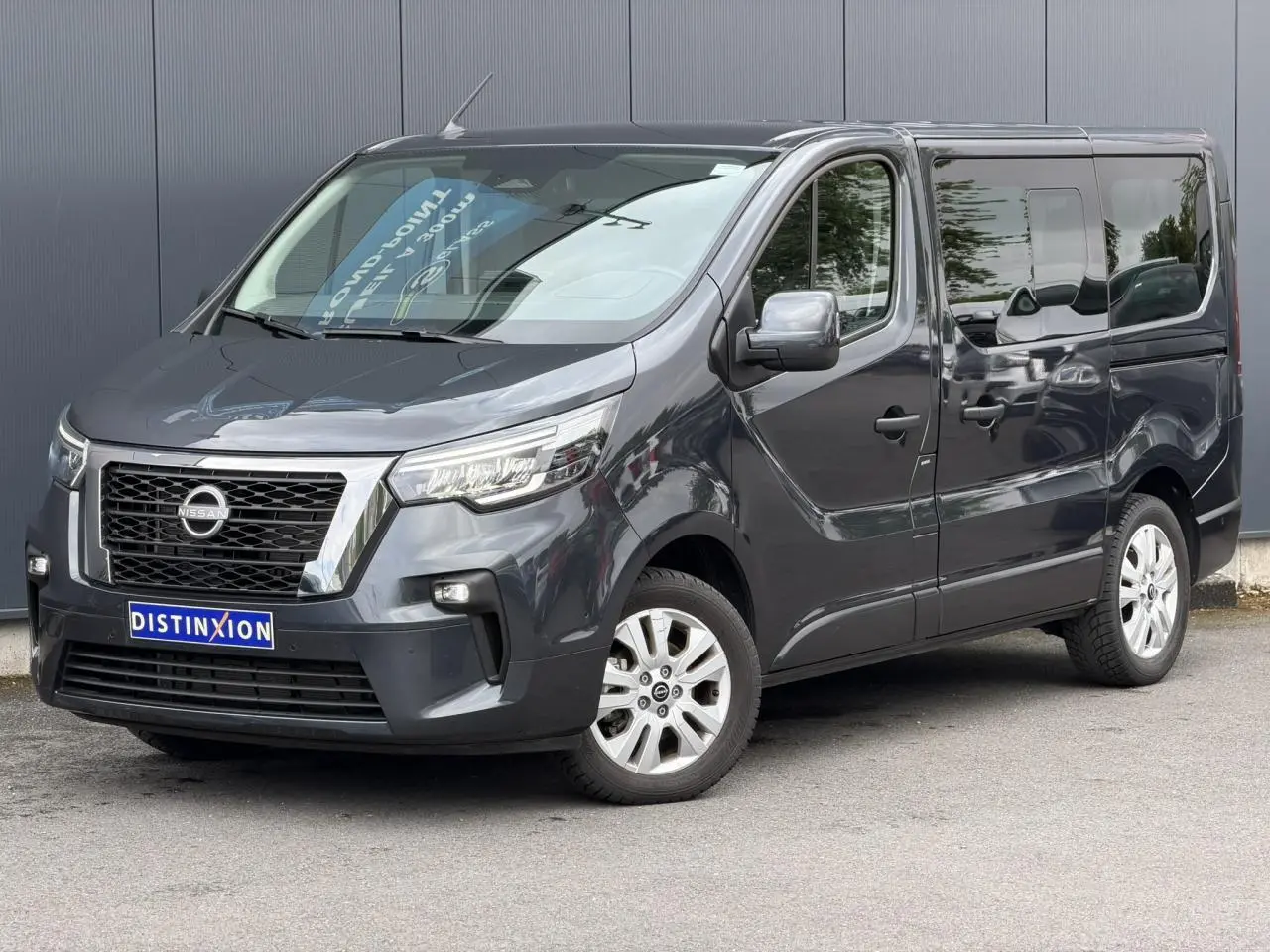 Nissan Primastar gris comète en vue 3/4 avant droit, avec hayon vitré et vitres latérales teintées.