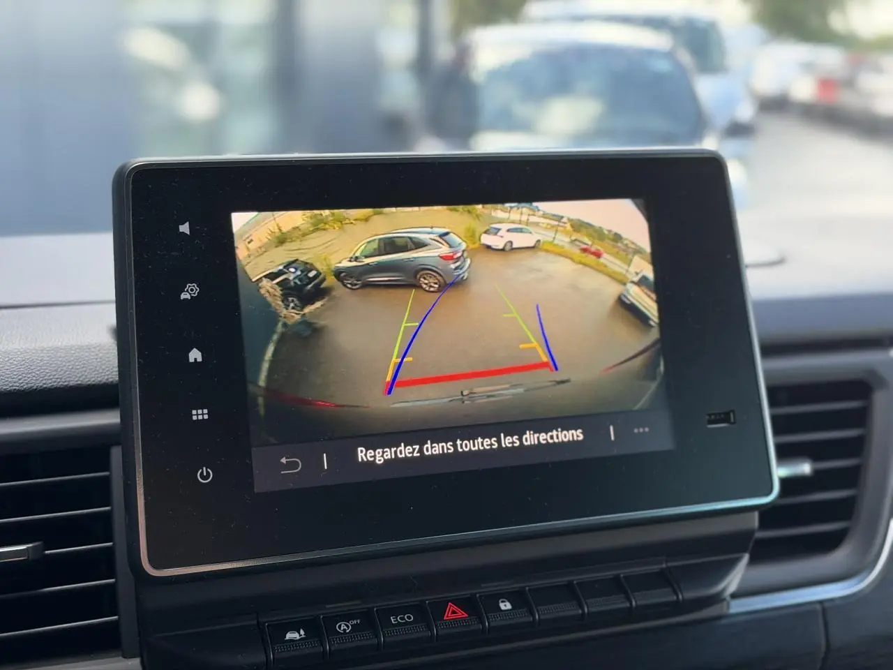 Écran de caméra de recul affichant un parking avec plusieurs véhicules, vu depuis l’habitacle du Nissan Primastar gris.
