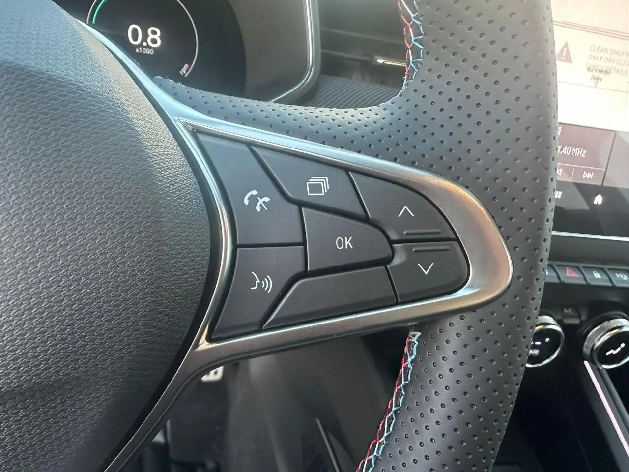 Gros plan sur les commandes multifonctions à gauche du volant perforé noir avec surpiqûres tricolores d’une Renault Clio V rouge.