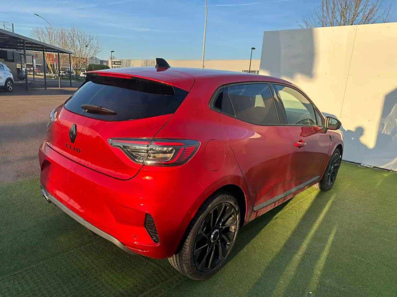 Vue 3/4 arrière droite d'une Renault Clio rouge foncé avec jantes noires et vitres teintées sur parking extérieur.