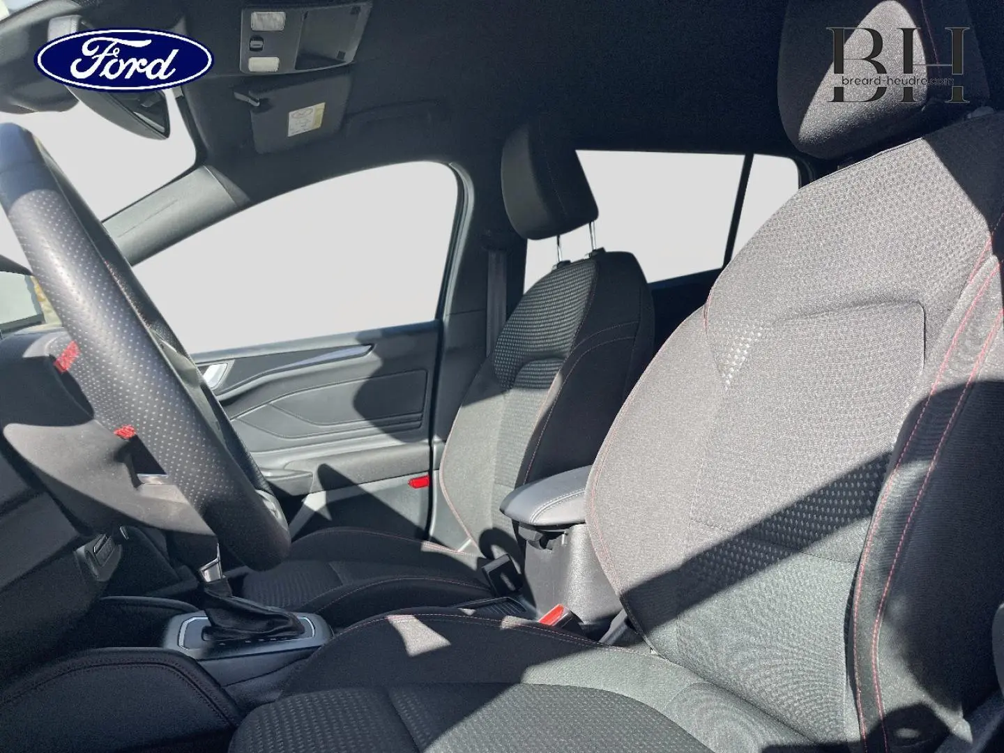 Intérieur avant de la Ford Focus SW 2024, sièges tissu noir avec surpiqûres rouges et volant cuir perforé.