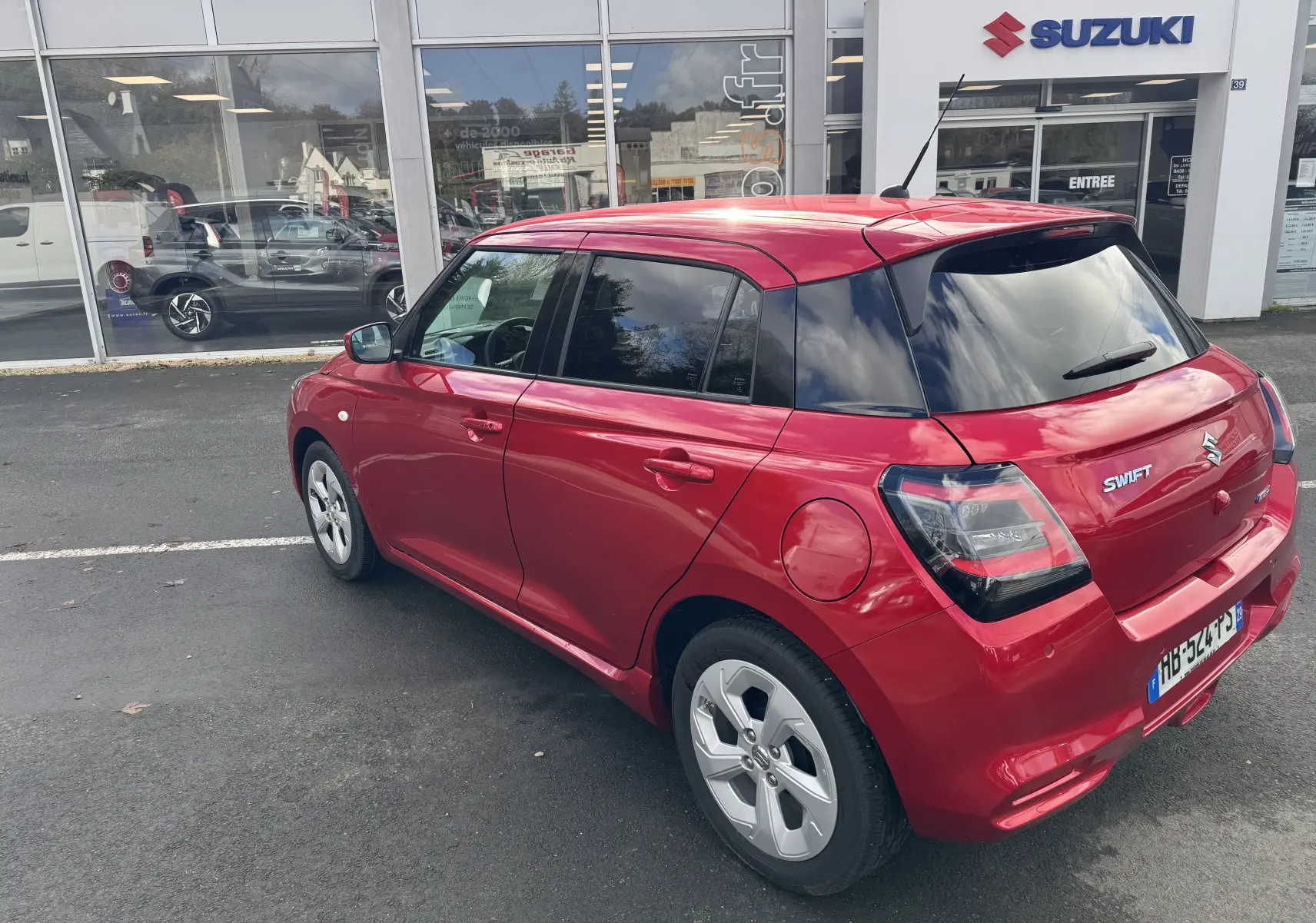 Vue 3/4 arrière droite d'une Suzuki Swift 1.2 Hybrid Privilege 2025 en rouge Burning Pearl, stationnée devant un concessionnaire.