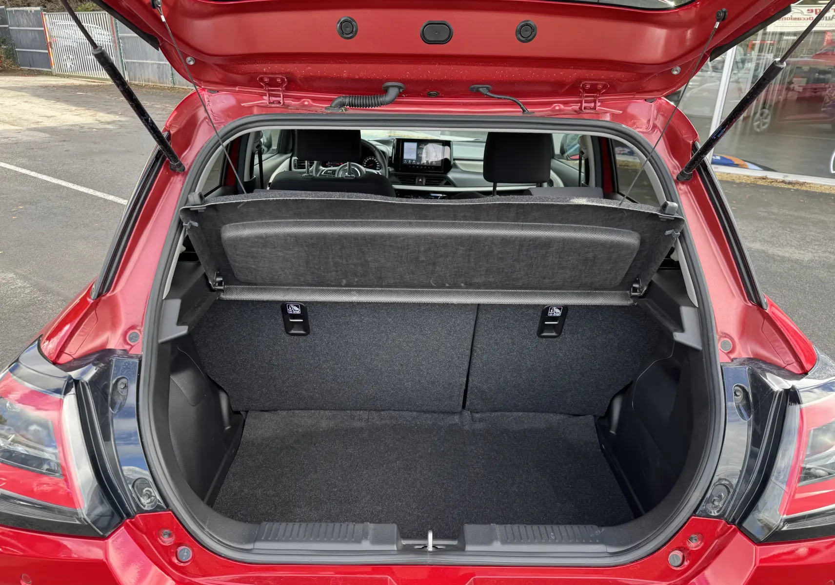 Coffre ouvert vu de l'arrière d'une Suzuki Swift rouge Burning Red Pearl Metal, intérieur noir et tableau de bord visible.