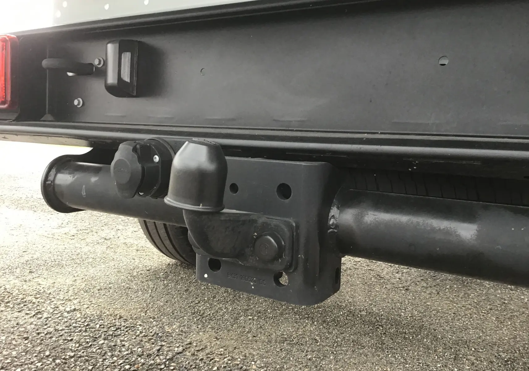 Gros plan sur l'attelage noir fixé sous le pare-chocs arrière du Ford Transit blanc, avec prise électrique visible.