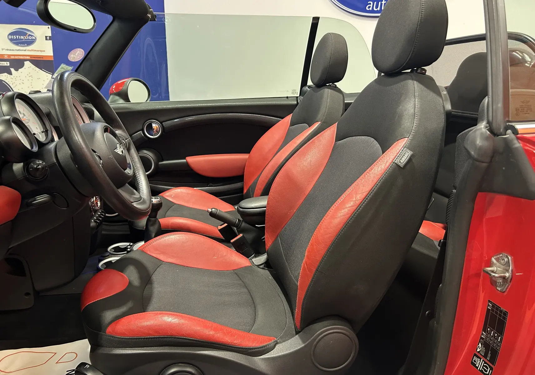 Intérieur côté gauche d'une MINI Cabriolet Cooper S rouge de 2012, sièges cuir rouge et noir, volant et levier manuel visibles.