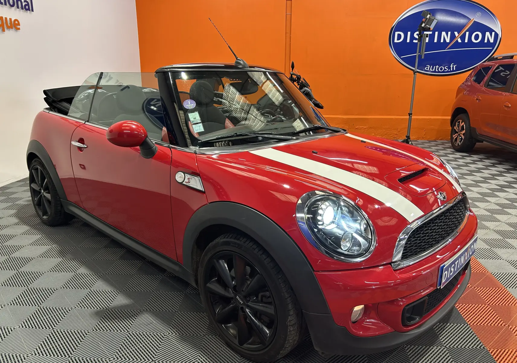 Vue 3/4 avant droite d'une MINI Cabriolet Cooper S rouge avec bandes blanches et jantes noires, capote ouverte.