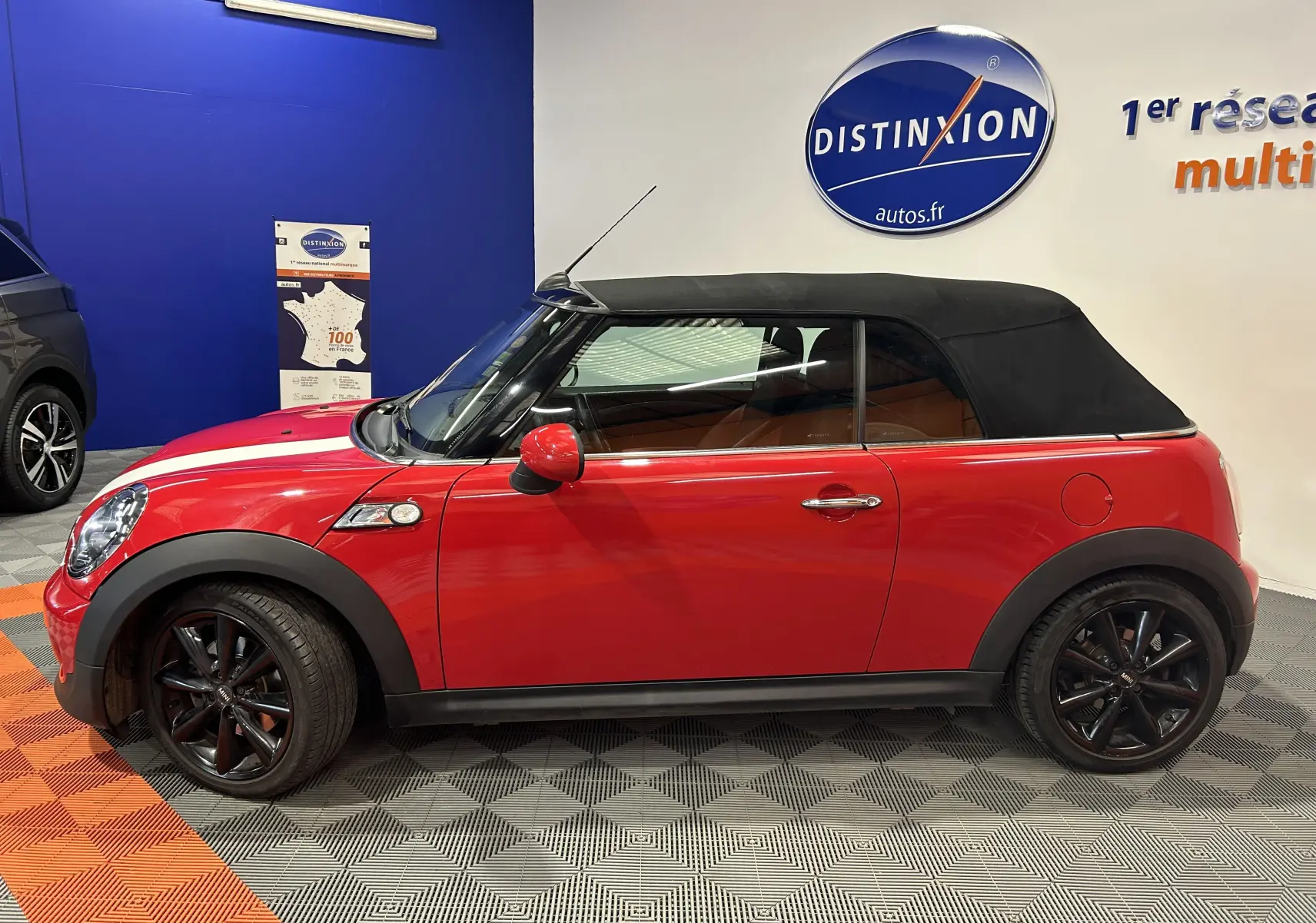 Profil droit d'une MINI Cabriolet Cooper S rouge avec capote noire et jantes noires en intérieur showroom.