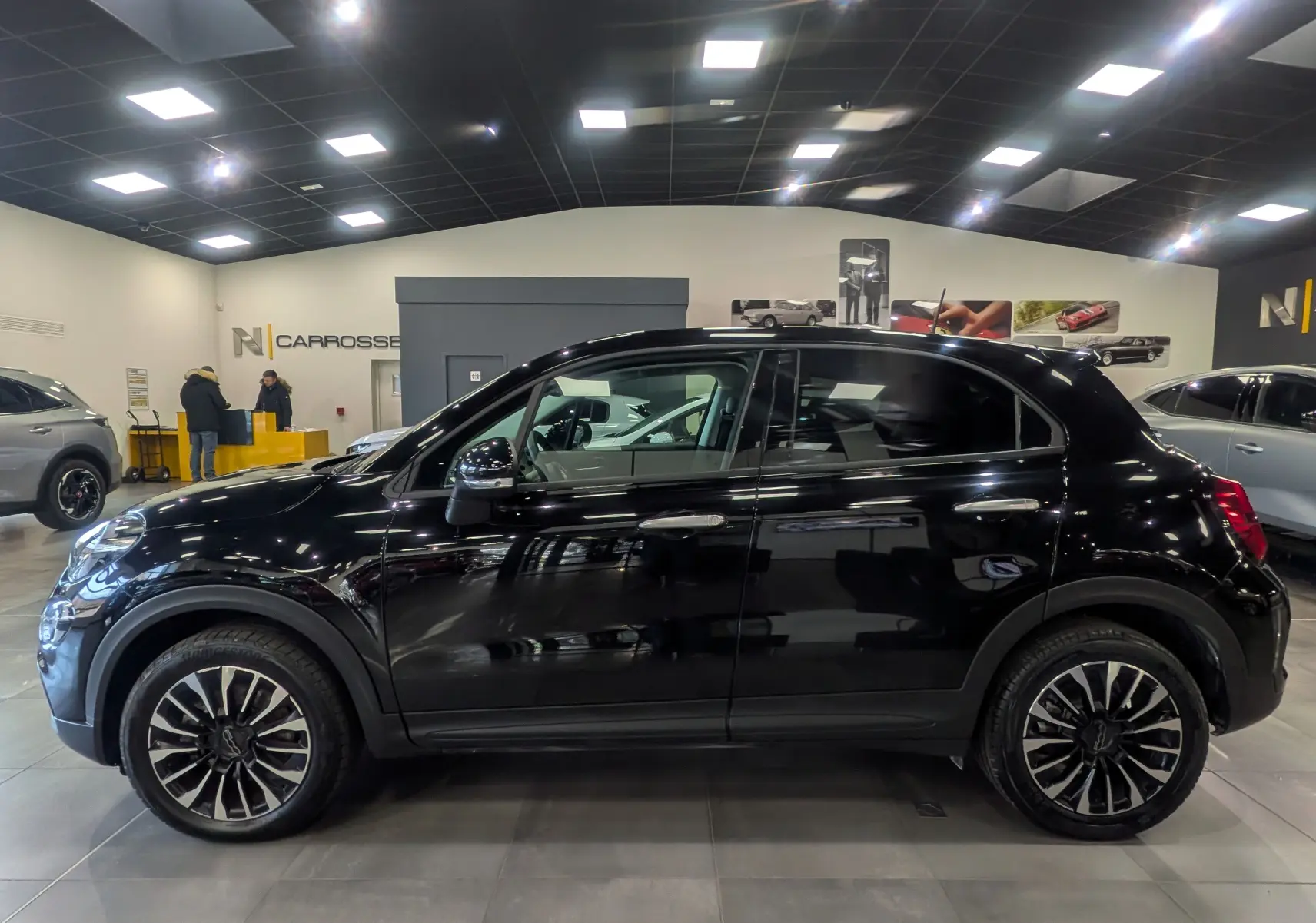 Vue de profil côté gauche d'une FIAT 500X 2024 noire Cinéma avec jantes alliage bicolores en intérieur showroom.