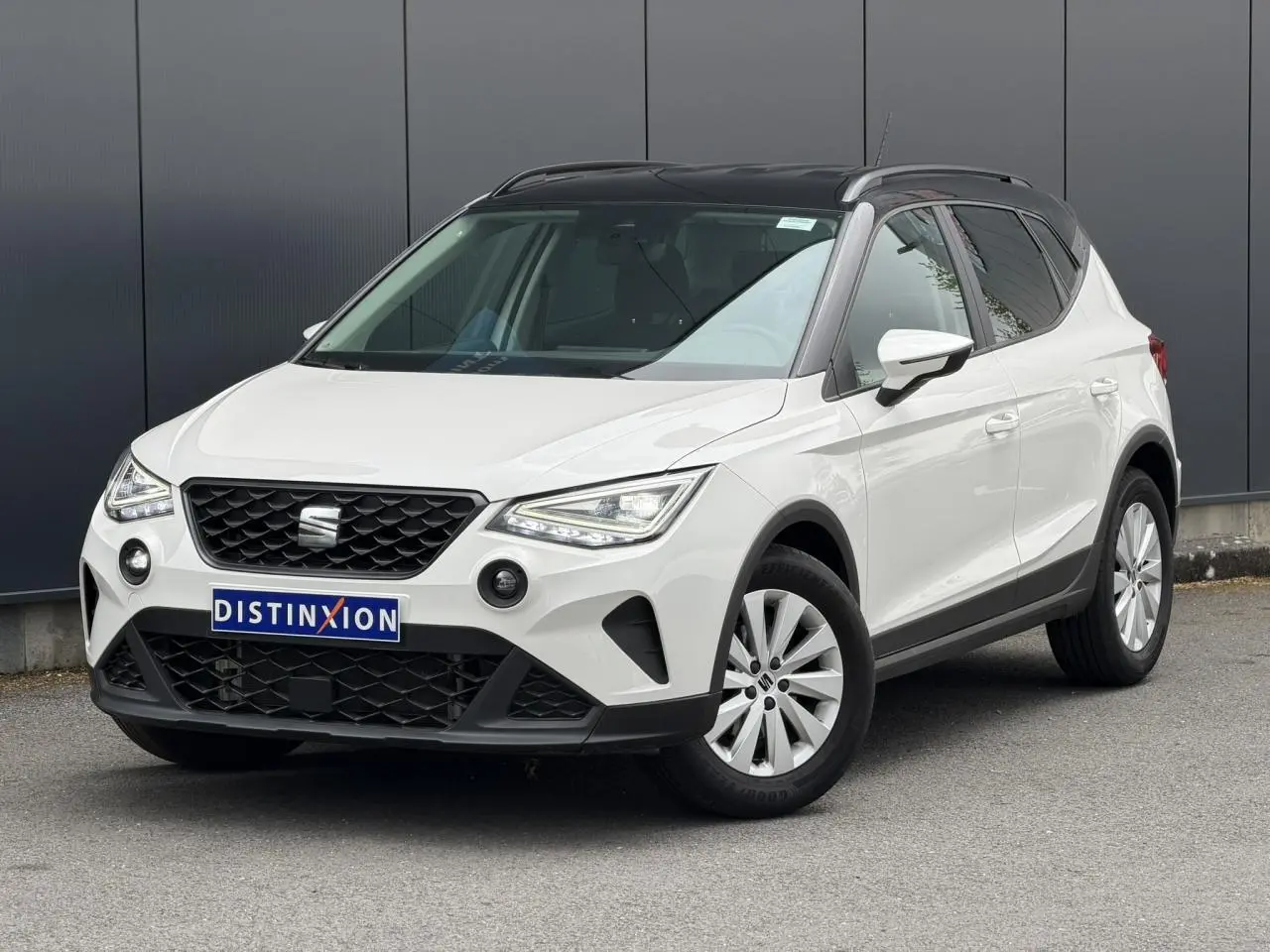 Vue 3/4 avant droite d'un SEAT Arona blanc avec toit noir et jantes alliage 16 pouces, garé devant un mur gris.