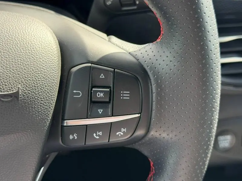 Gros plan sur les commandes multifonctions noires du volant cuir perforé avec surpiqûres rouges de la Ford Focus gris lunaire.