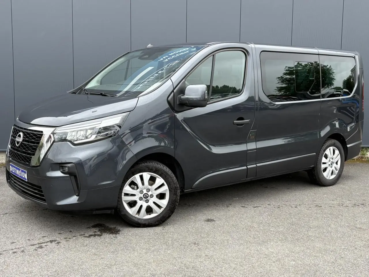Nissan Primastar gris comète vue 3/4 avant droit, hayon vitré et jantes alliage 17 pouces visibles.