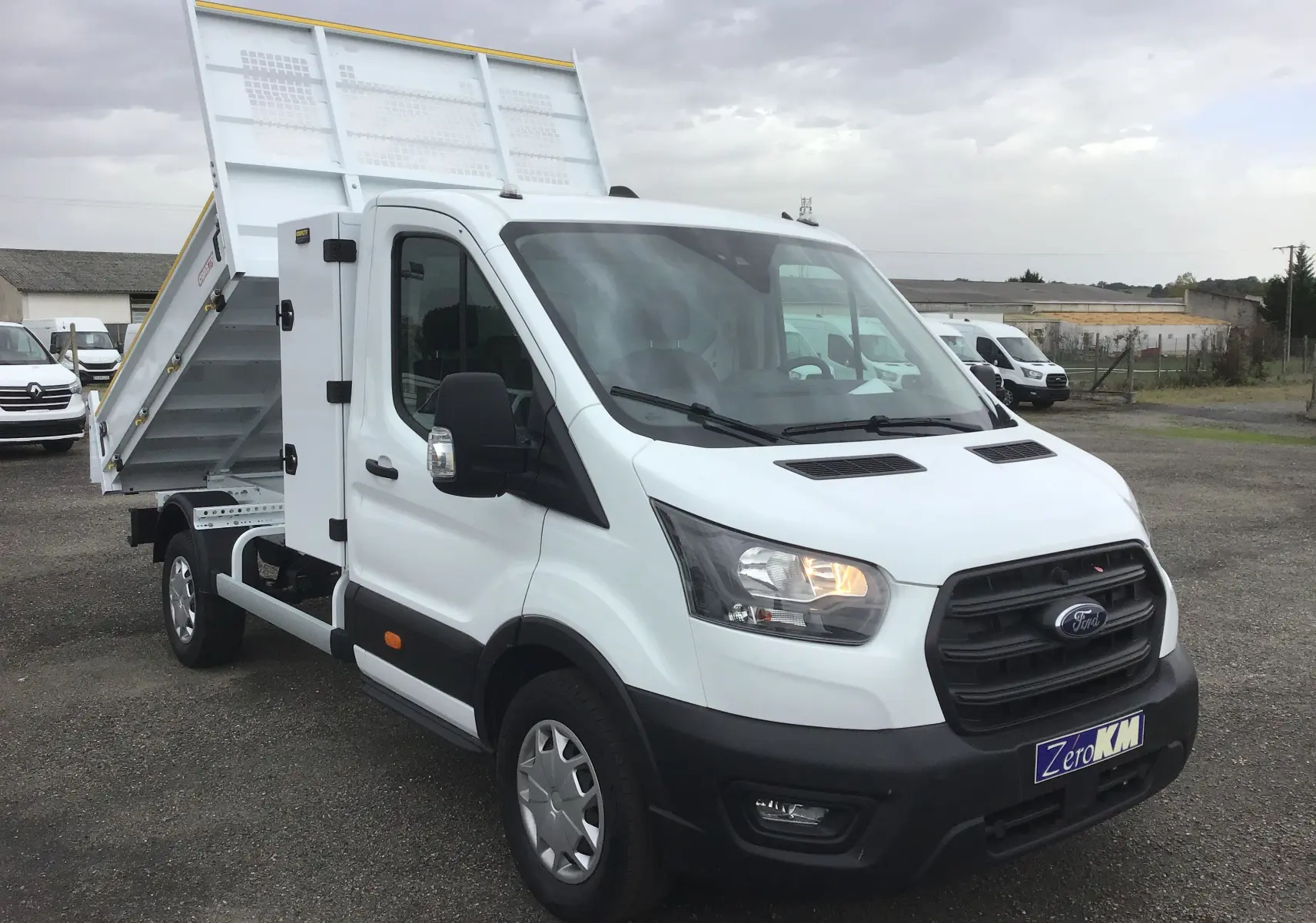 Vue 3/4 avant droit d'un Ford Transit blanc avec benne relevée et coffre latéral acier sur parking extérieur.