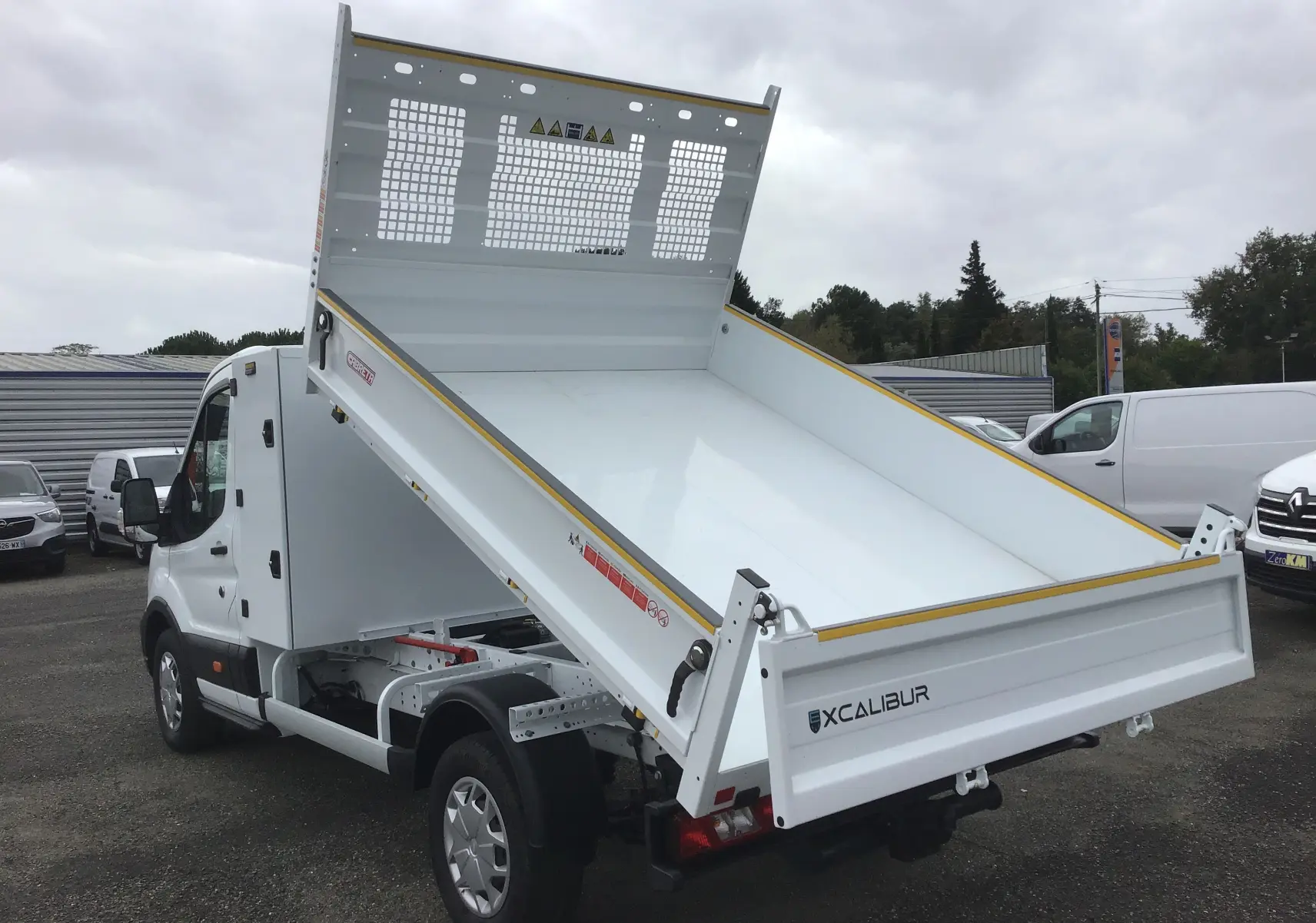 Vue 3/4 arrière droite d'un Ford Transit Chassis Cabine blanc avec benne relevée et coffre acier sur un parking.
