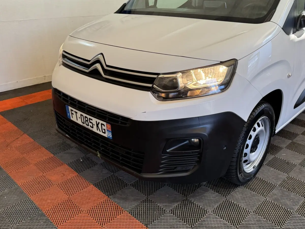 Vue 3/4 avant droit d'un Citroën Berlingo blanc avec pare-chocs noir et phares allumés en intérieur.