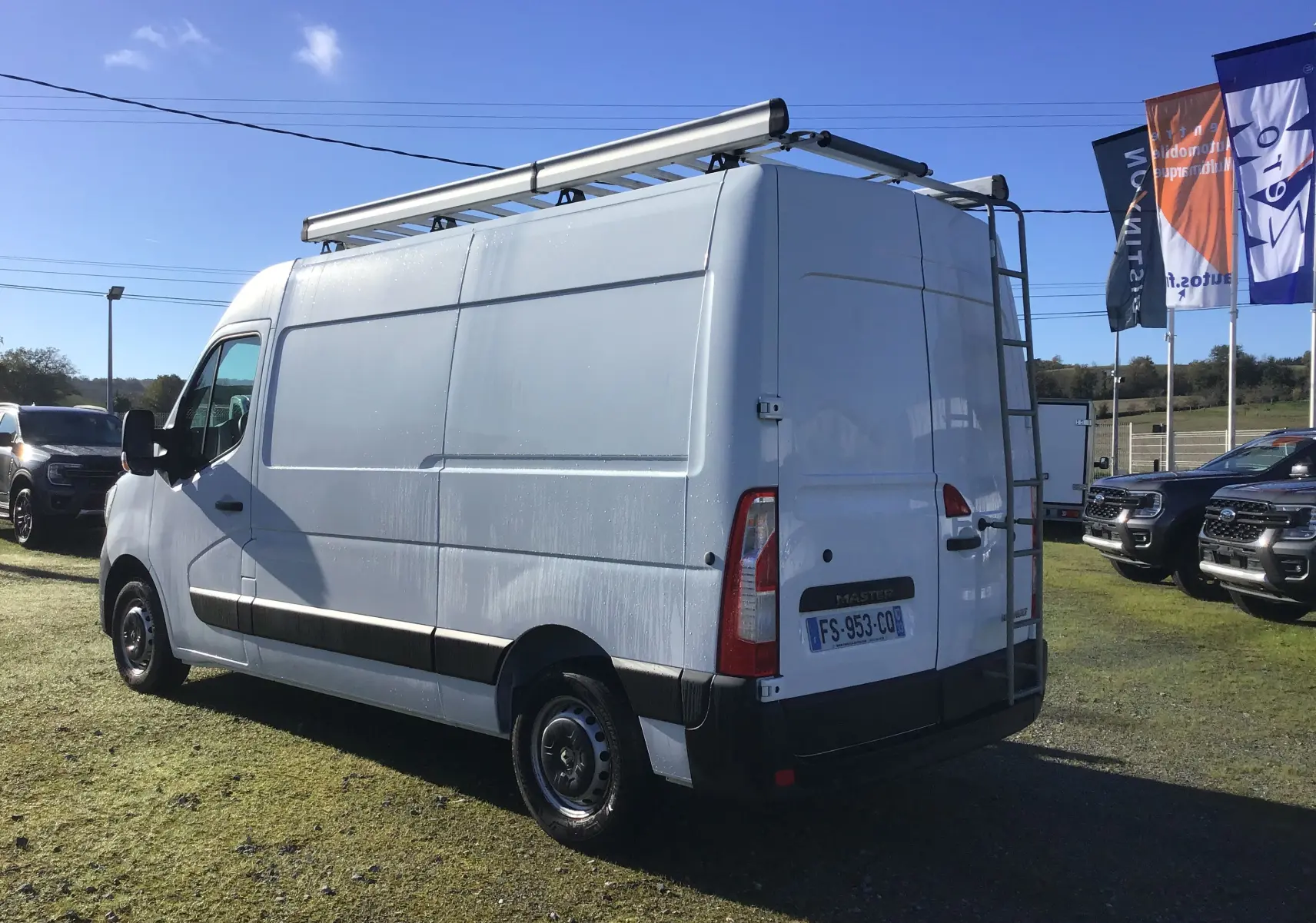 Vue 3/4 arrière droite d'un utilitaire Renault Master blanc avec galerie de toit et échelle arrière métallique.