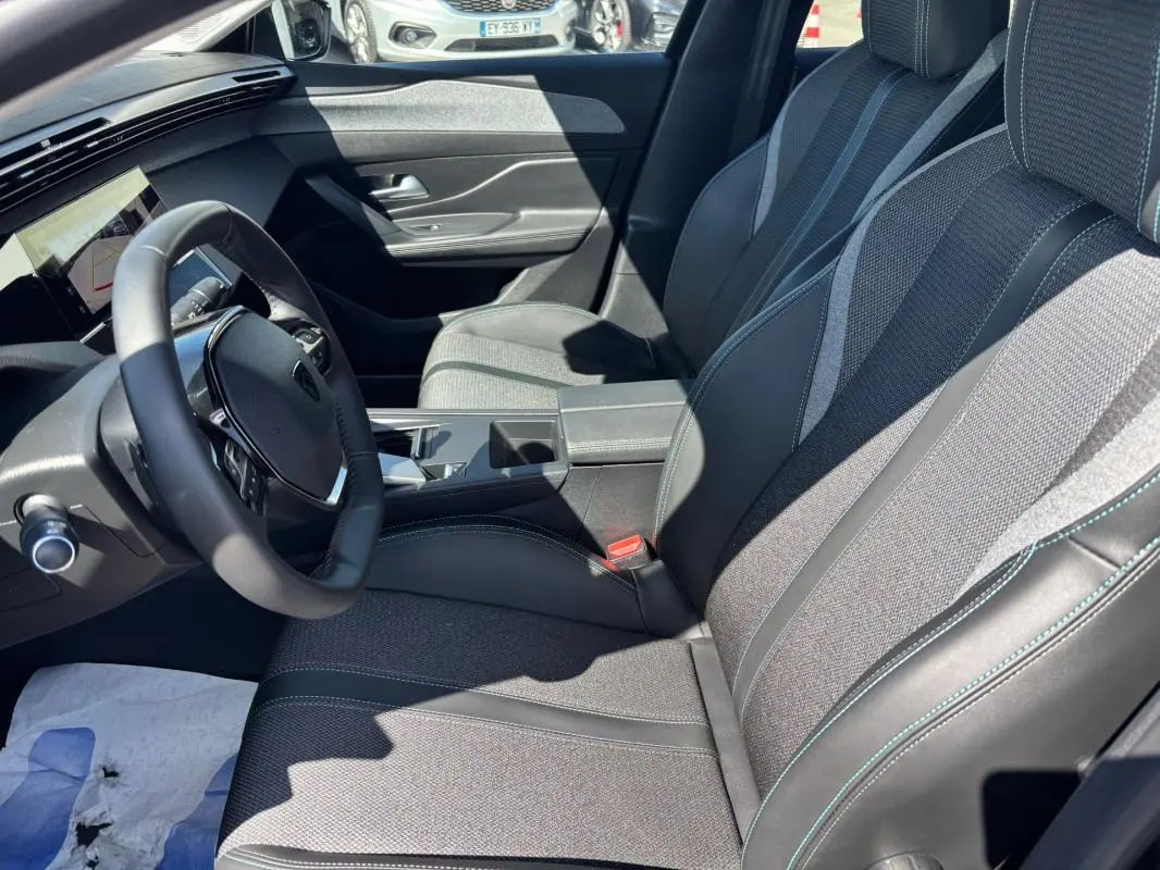 Vue intérieure côté conducteur de la Peugeot 408 Hybrid 2025 noire, avec sièges tissu et cuir, accoudoir central et volant multifonction.
