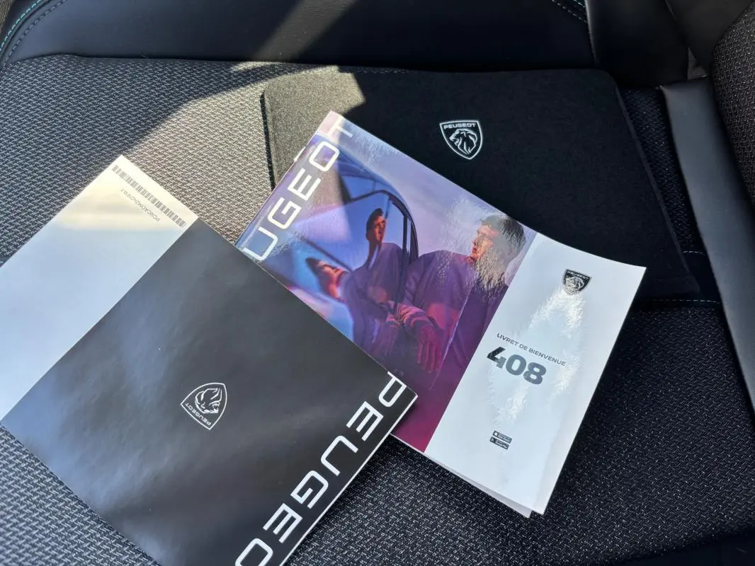 Vue rapprochée des brochures Peugeot 408 posées sur un siège tissu noir à motifs, avec logo Peugeot visible.