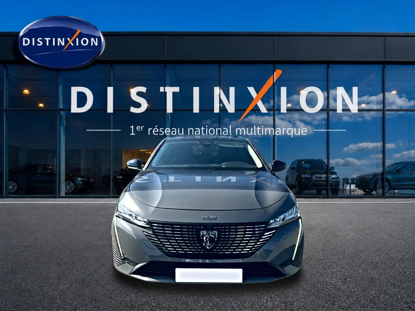 Peugeot 308 gris 3/4 avant face, avec calandre distinctive et phares LED allumés devant showroom Distinxion.
