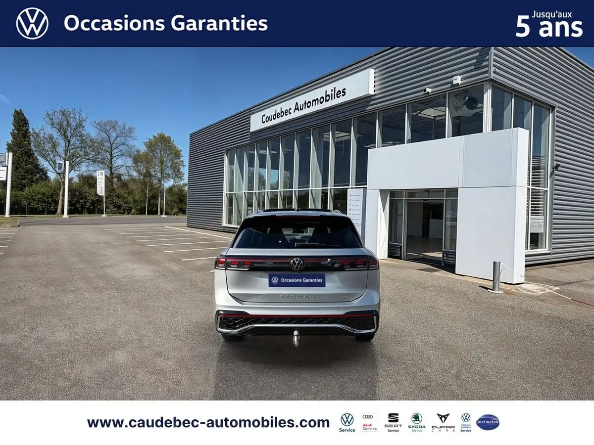 Vue arrière d’un Volkswagen Tiguan eHybrid 2025 argent Dolomite avec hayon électrique et crochet d’attelage escamotable, devant un bâtiment.