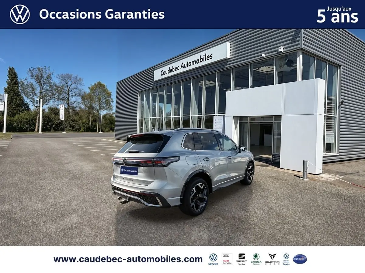Vue 3/4 arrière droite d’un Volkswagen Tiguan eHybrid argent Dolomite avec jantes 19 pouces devant un showroom Caudebec Automobiles.