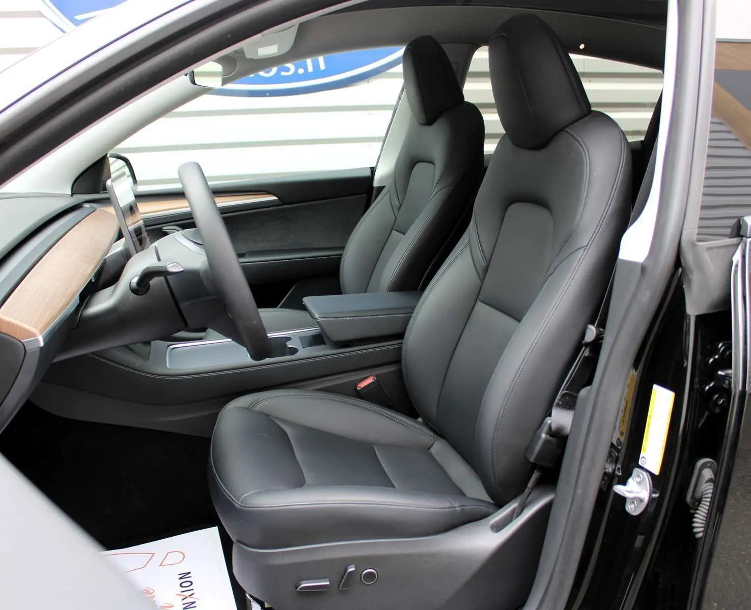 Intérieur de la Tesla Model Y 2024 en cuir noir, vue côté conducteur avec tableau de bord bois et écran central.