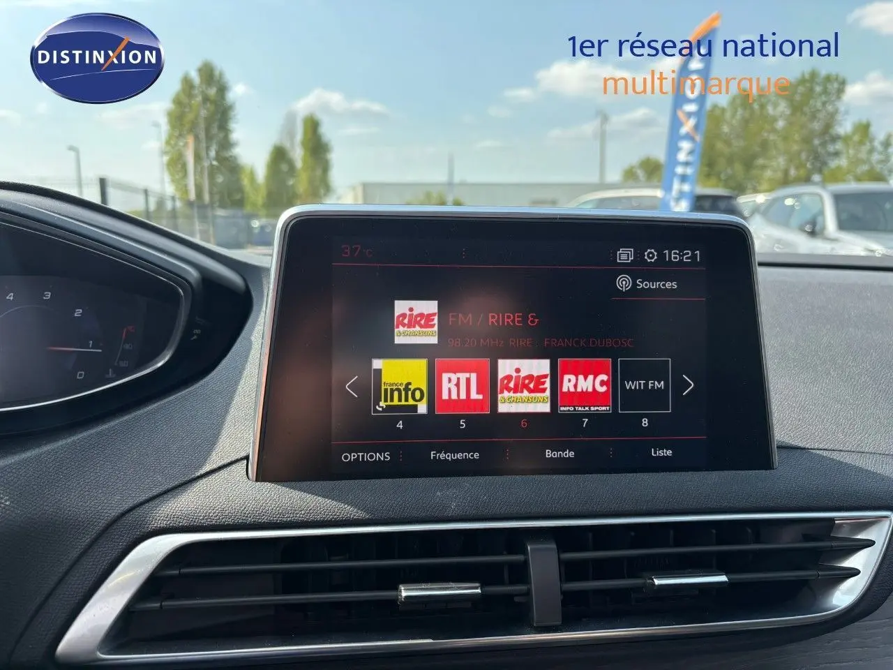 Écran multimédia central du Peugeot 3008 GT blanc nacré, avec tableau de bord partiel et vue extérieure floutée.