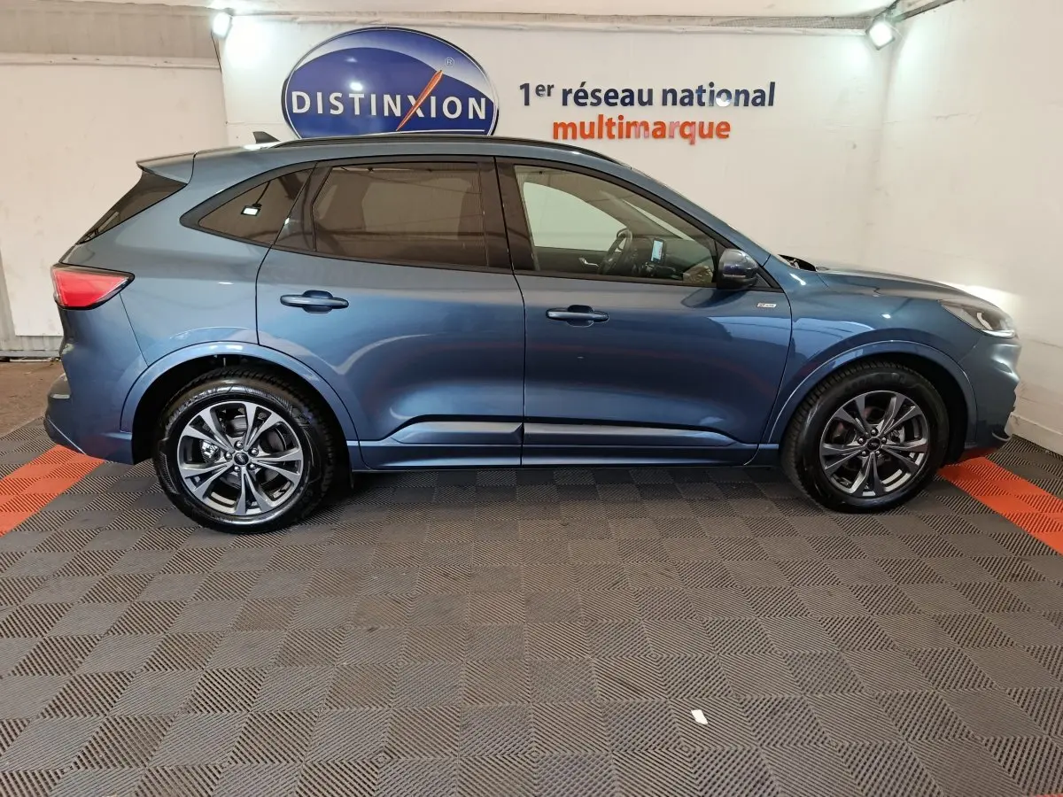 Ford Kuga 2024 bleu en profil côté gauche, avec jantes alliage et vitres teintées dans un showroom.