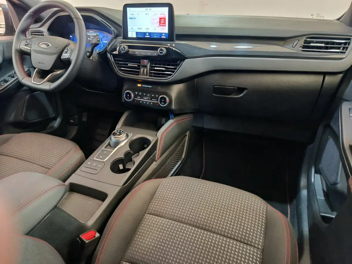Intérieur du Ford Kuga 2024 St Line, vue côté conducteur, tableau de bord noir avec écran tactile et sièges tissu gris à surpiqûres rouges.