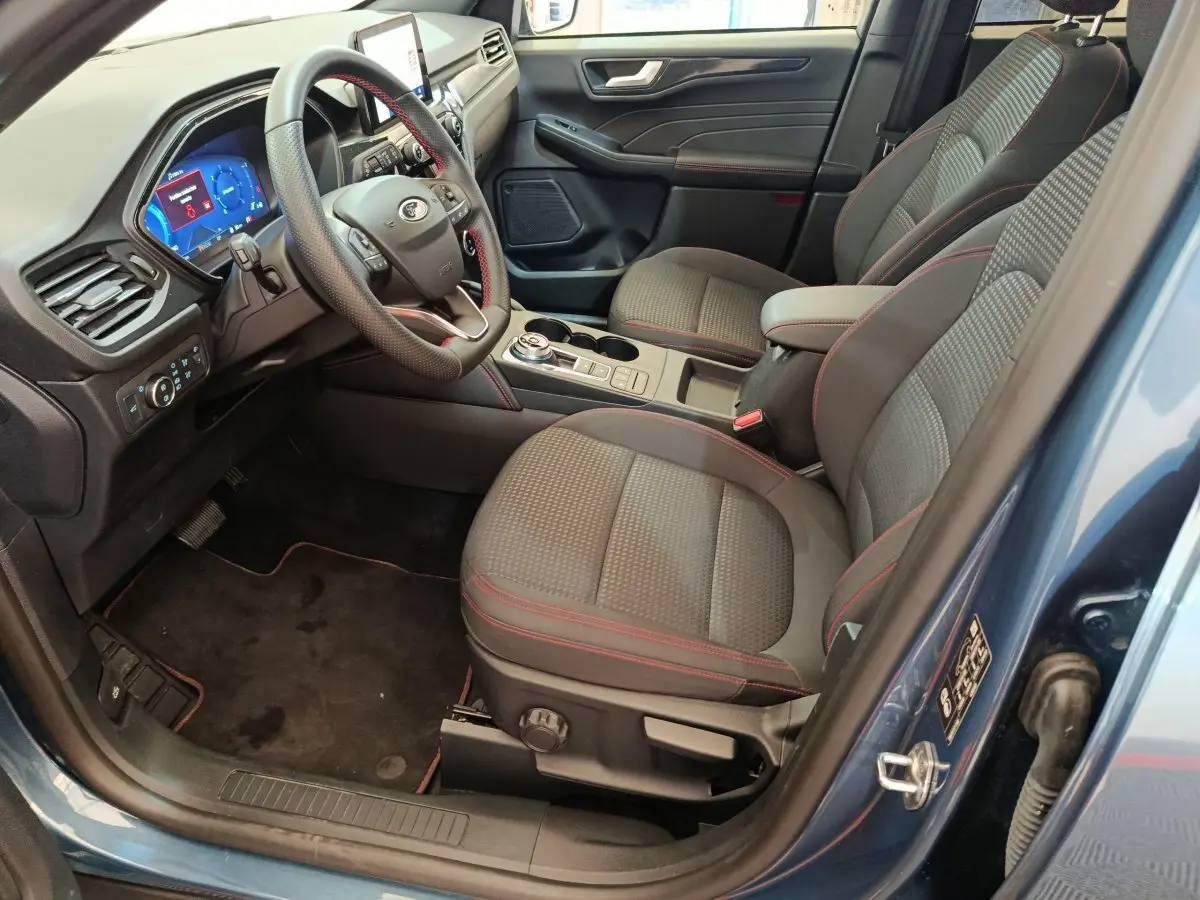 Vue intérieure côté conducteur du Ford Kuga bleu 2024, sièges tissu gris avec surpiqûres rouges et tableau de bord numérique.