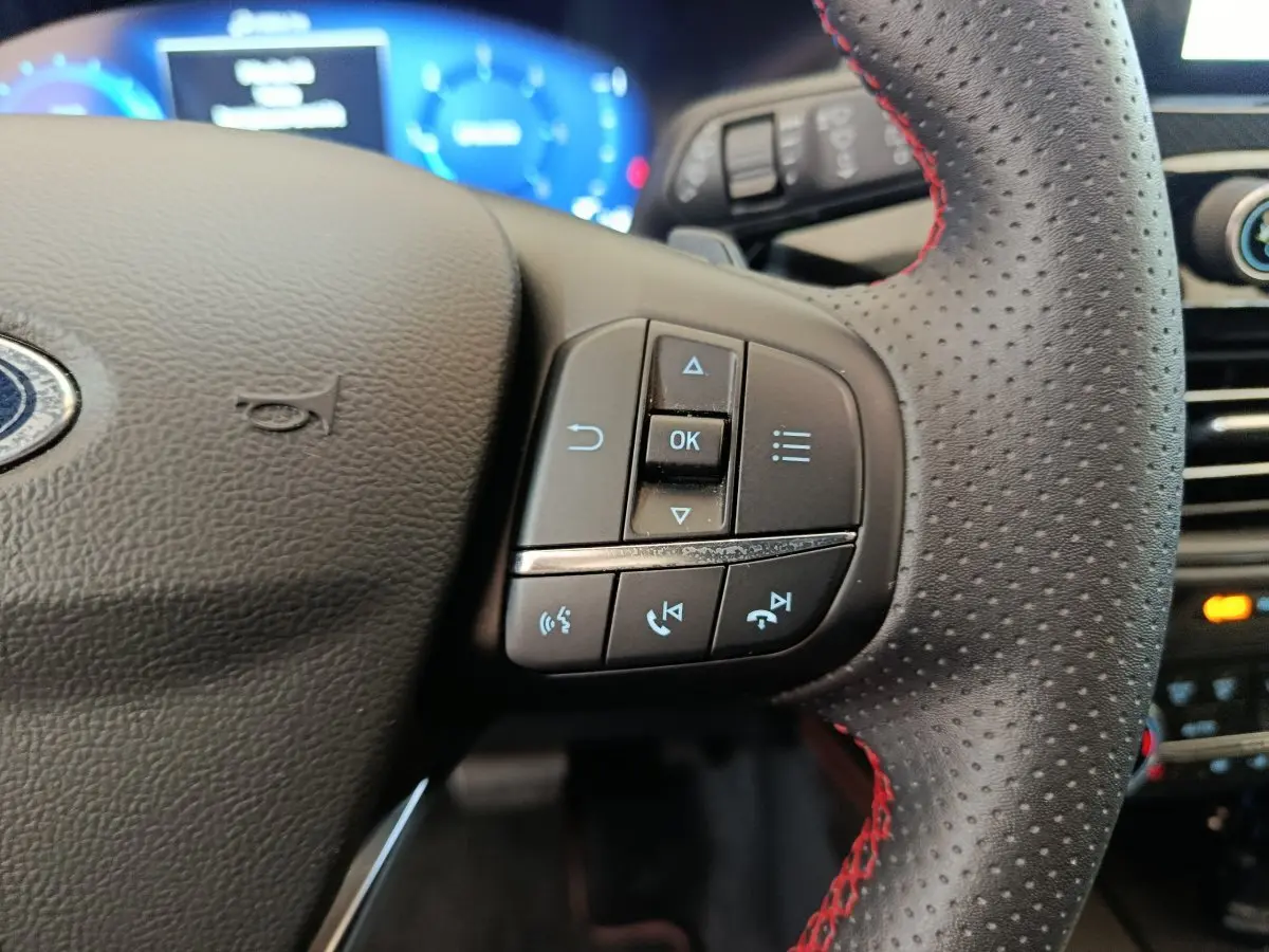 Gros plan sur le volant en cuir noir perforé avec surpiqûres rouges du Ford Kuga 2024, boutons multifonctions visibles.