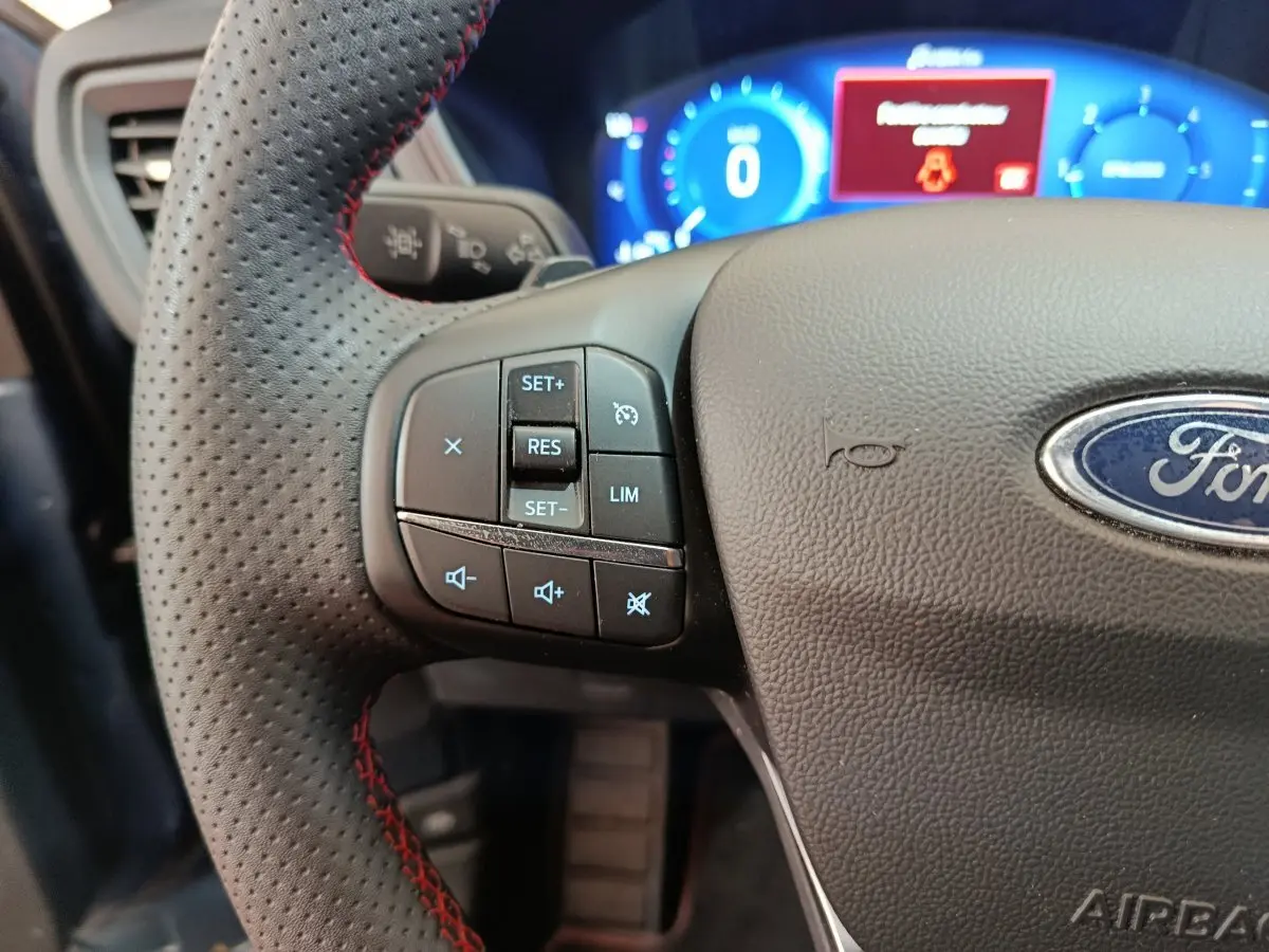 Gros plan sur le volant perforé avec surpiqûres rouges du Ford Kuga 2024, tableau de bord numérique bleu en arrière-plan.