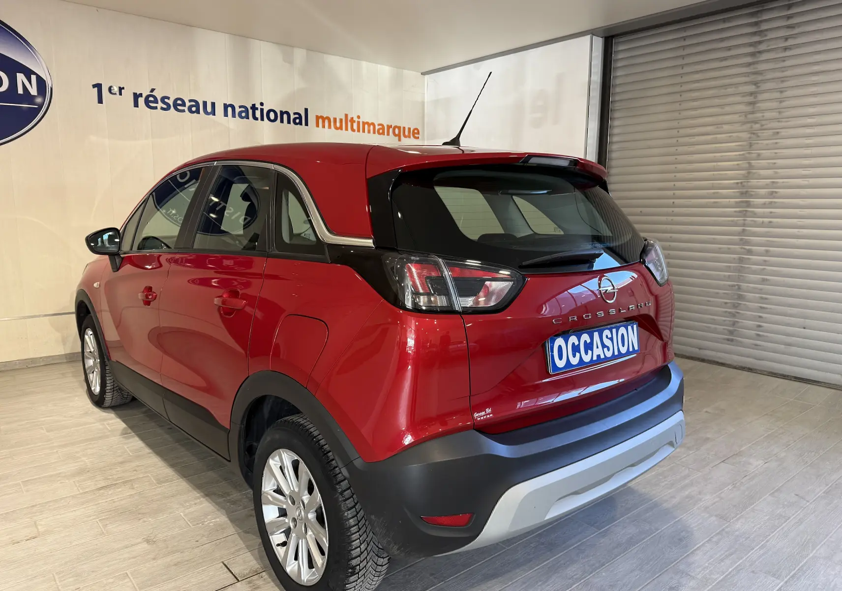 Vue 3/4 arrière droite d’un Opel Crossland rouge 2022 avec badge Crossland et plaque "OCCASION" en intérieur showroom.