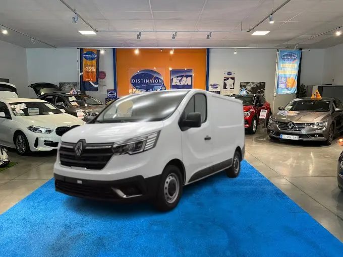 Renault Trafic Fourgon blanc en 3/4 avant droit, présenté en showroom sur tapis bleu avec autres véhicules autour.