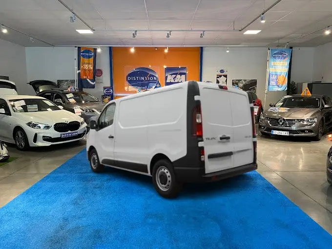 Vue 3/4 arrière d’un Renault Trafic Fourgon blanc L1H1 dans un showroom avec sol bleu et autres véhicules autour.
